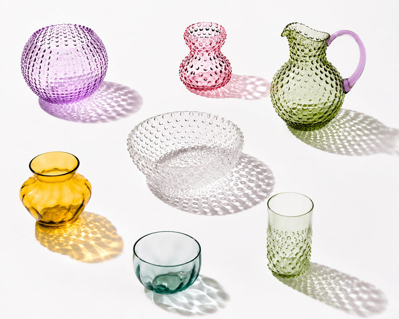 Colorful mouth-blown Anna von Lipa glasses and jugs in citron, pink and amber on a white table in bright sunlight, casting long shadows.