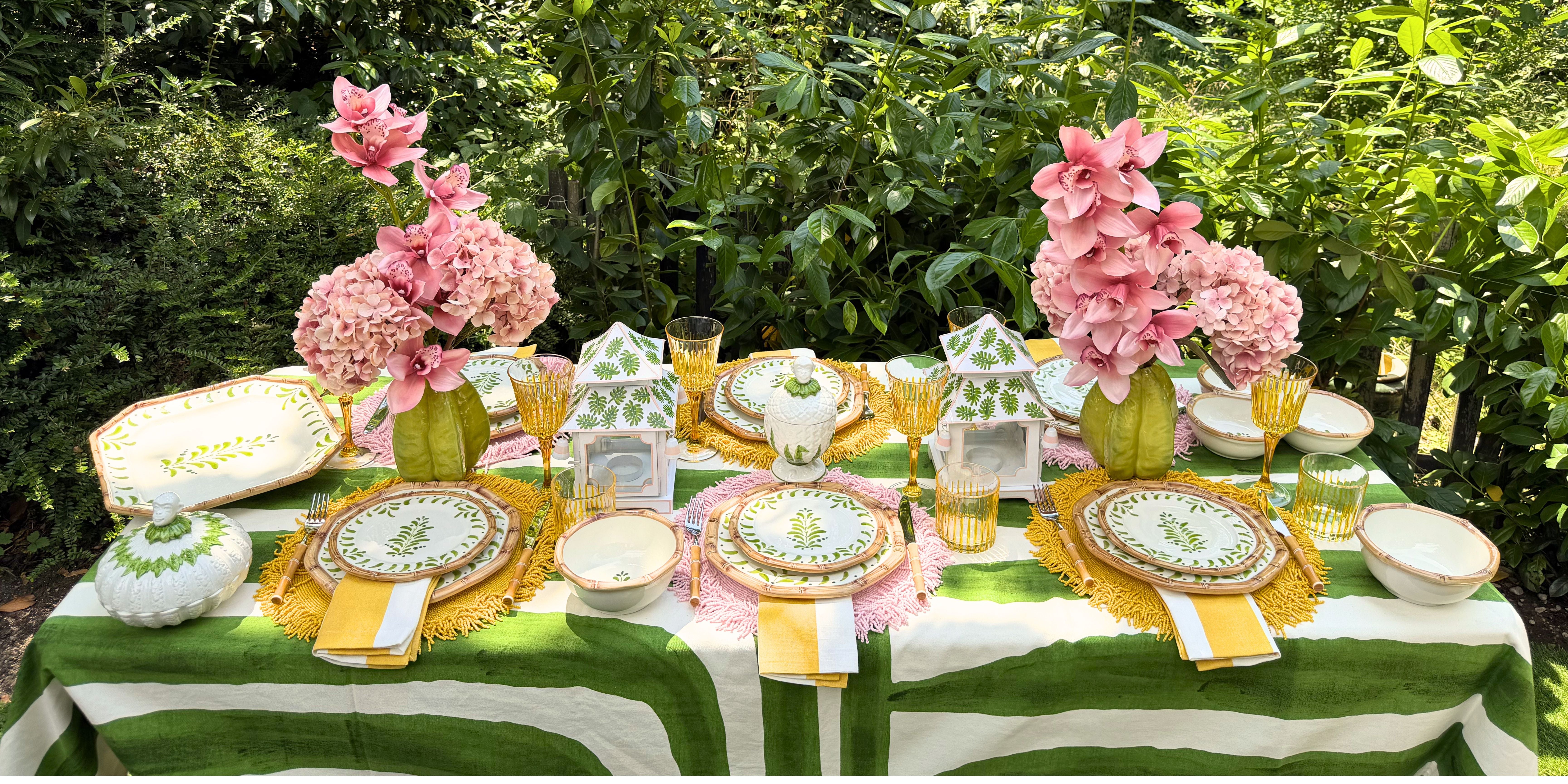 An alfresco luxury tablescape from The Eden Collection by Table Desires x Les Ottomans, featuring green hand-painted tableware, a striped tablecloth, yellow and pink placemats, and lush pink orchids in an outdoor garden setting.