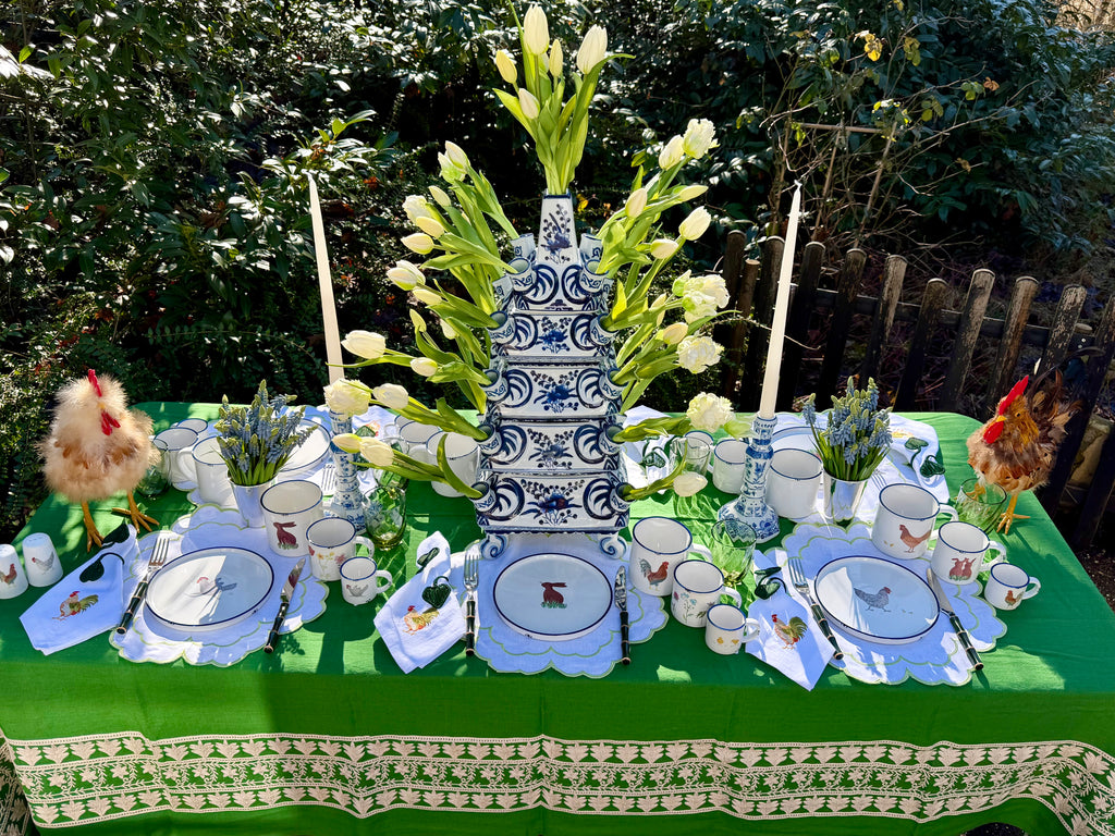 Frühlingshaft gedeckter Ostertisch im Freien mit grüner Tischdecke, weiß-blauem Etagen-Blumenhalter, Tulpen und Geschirr mit Hühner- und Hasen-Motiven.